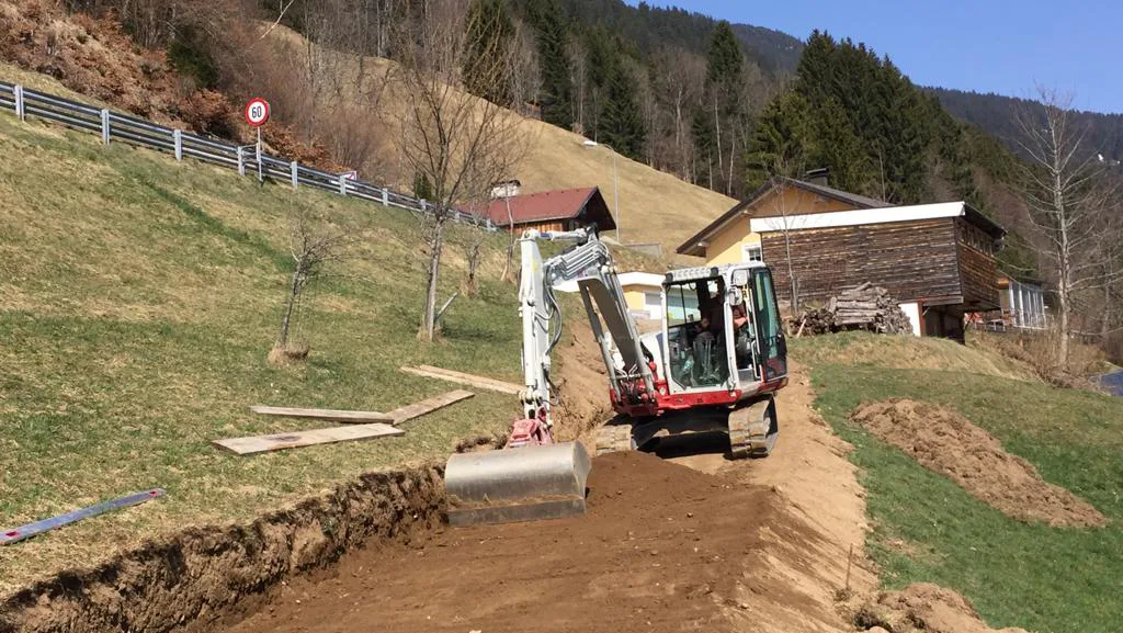 Bagger beim Aushub an einem steilen Hang, im Hintergrund Häuser einer Montafoner Streusiedlung