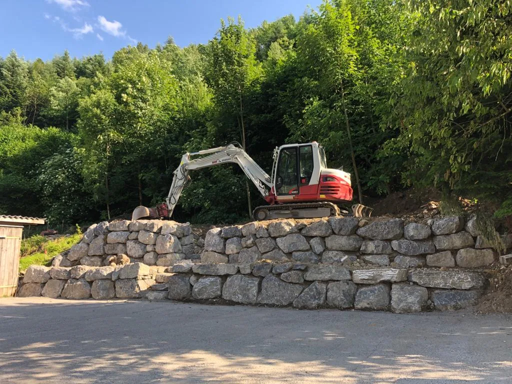 Fertige Natursteinmauer aus großen Blöcken, oben darauf ein Bagger am Waldrand
