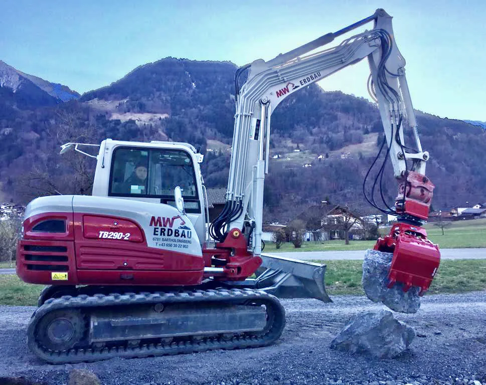 Bagger Takeuchi TB290-2 mit der Aufschrift MW Erdbau, 6781 Bartholomäberg, hält einen Stein im Sortiergreifer