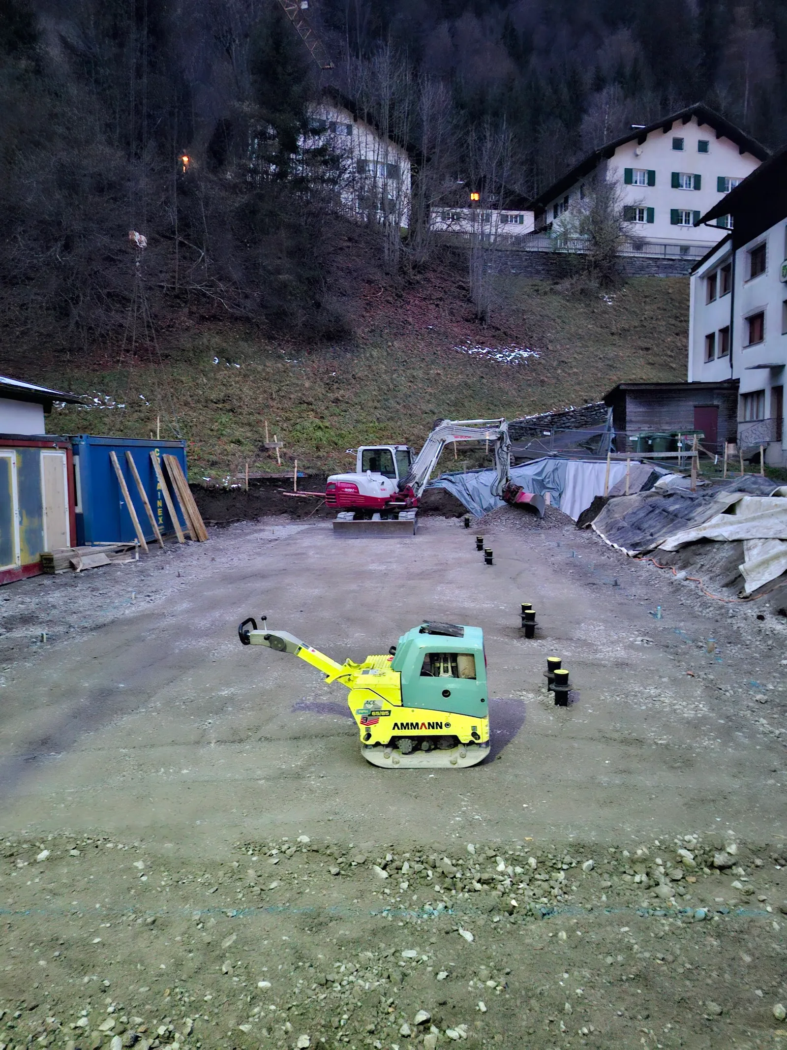 Baustelle in der Dämmerung: Rüttelplatte auf verdichtetem Schotter, dahinter ein Bagger und gesetzte Fundamentpunkte