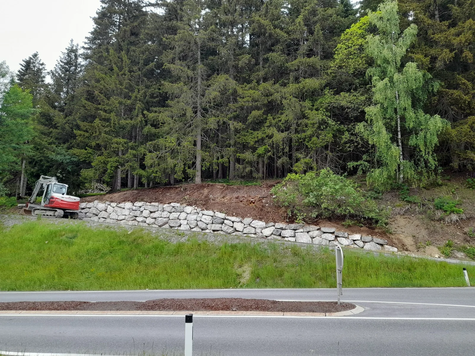 Neu gesetzte Natursteinmauer entlang einer Straße, daneben ein Bagger vor einem Fichtenwald