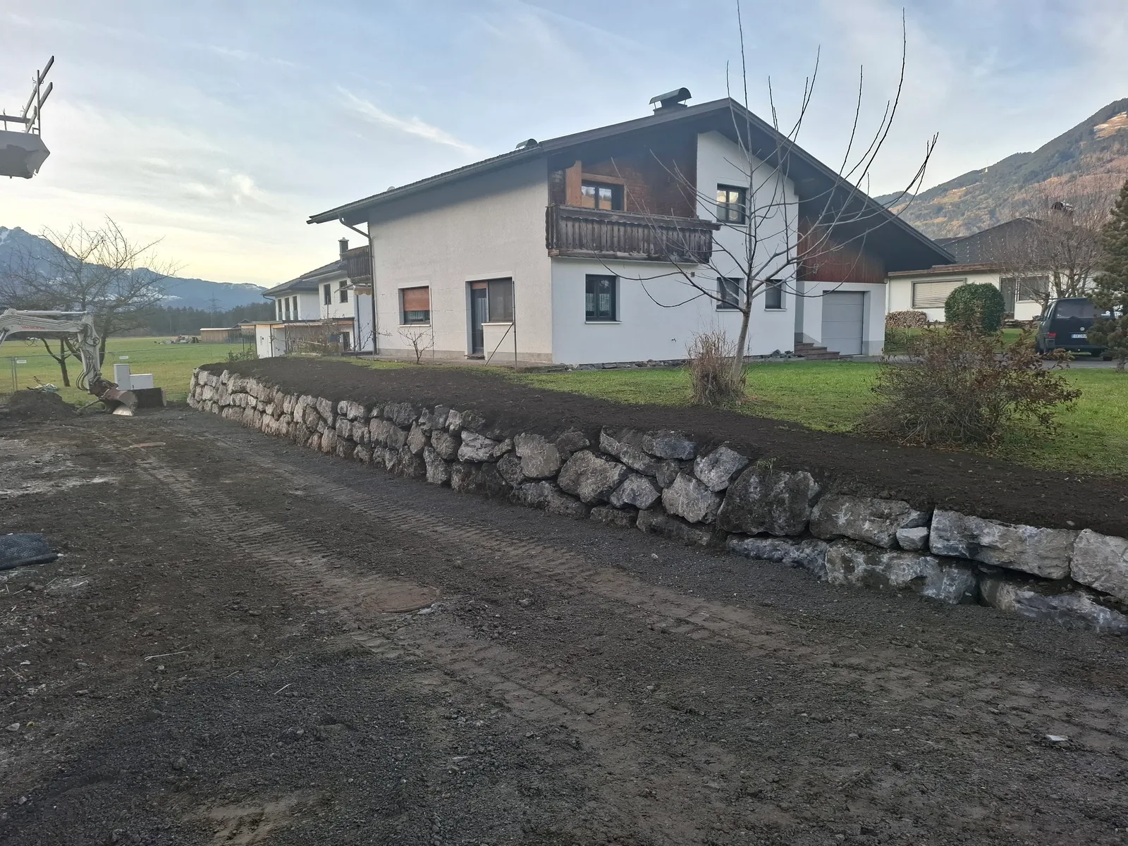 Fertige Natursteinmauer und frisch humusierte Böschung vor einem Wohnhaus, dahinter die Berge