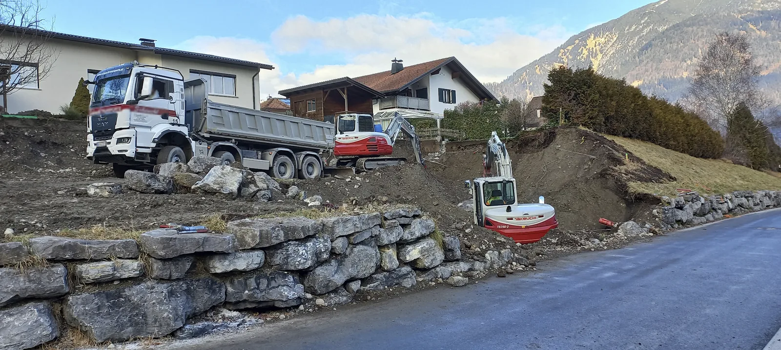 Baustelle an einer Straße mit Kipper, zwei Baggern und einer gesetzten Steinmauer